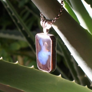 Peut inclure: Un collier pendentif rectangulaire avec une pierre d'opale marron et bleue. Le pendentif est serti dans un cadre en métal argenté et est suspendu à une chaîne délicate. Le fond présente des feuilles d'aloe vera vertes.