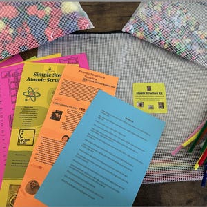 May include: An educational kit with colourful elements. The image shows printed materials, including a periodic table and atomic structure guides, along with bags of beads and pipe cleaners. The kit is designed for learning about atomic structure.