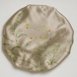 May include: A beige satin sleep cap with a floral print featuring green leaves and small yellow and orange flowers.