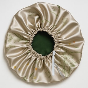 Op de afbeelding: Een beige satijnen slaapmuts met een bloemenprint en een wit elastiek. De muts is bovenaan gerimpeld en heeft een groene voering.