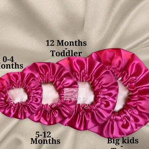 May include: Three pink satin hair bonnets in different sizes. The smallest bonnet is labeled "0-4 Months", the middle bonnet is labeled "5-12 Months", and the largest bonnet is labeled "12 Months Toddler".