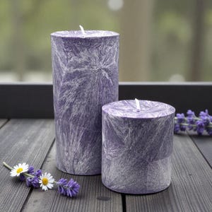 May include: Two purple pillar candles with a textured, crystallized design. The candles are cylindrical, one taller than the other, and have white wicks. The candles are on a dark wooden surface, with sprigs of lavender and daisies.