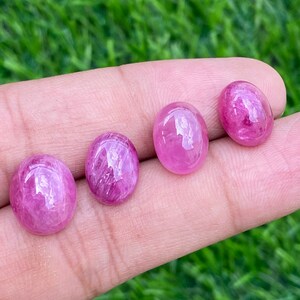 May include: Four pink oval-shaped gemstones, likely rubies, are displayed on a finger. The gemstones are smooth and have a slight shimmer.