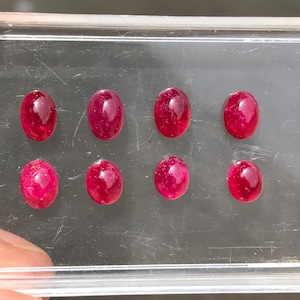 May include: Eight oval-shaped, deep red gemstones arranged in two rows of four inside a clear plastic display case. Each stone features a smooth, polished cabochon cut with a reflective surface, showcasing a consistent vibrant crimson color throughout.