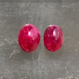 Könnte beinhalten: Zwei oval geformte, im Cabochon-Schliff geschliffene Edelsteine in einer tiefen, gesättigten magentaroten Farbe. Die Steine haben eine glatte, polierte Oberfläche mit sichtbaren inneren Einschlüssen und natürlichen Strukturvariationen.