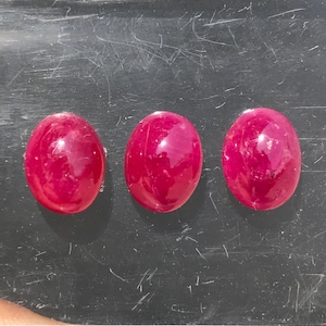 Puede incluir: Tres rubíes de forma ovalada, tallados en cabujón, con un color rojo intenso y vibrante y una superficie lisa y pulida. Las piedras preciosas están dispuestas en una fila horizontal, mostrando una calidad translúcida e inclusiones naturales.