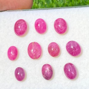 May include: Nine pink oval-shaped gemstones with a star-like pattern inside. The gemstones are arranged in a row on a white surface.