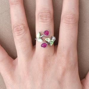 Könnte beinhalten: Ein silberner Ring mit einem Ast-Design, das mit grünen und rosafarbenen Edelsteinen besetzt ist. Der Ring befindet sich an einem Finger.