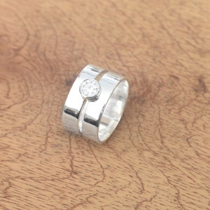Puede incluir: Un anillo de plata con una sola piedra transparente engastada en el centro. El anillo tiene una banda texturizada y una segunda banda que se envuelve alrededor de la parte superior del anillo.