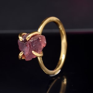 May include: A gold ring with a raw, red garnet gemstone set in the center. The ring is simple and elegant, with a smooth band.