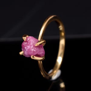 May include: A gold ring with a raw pink ruby gemstone. The ruby is set in a simple, modern design.