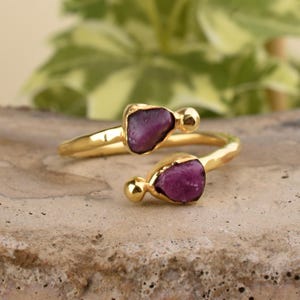 Puede incluir: Un anillo de oro con dos piedras preciosas púrpuras. El anillo es una banda simple con un acabado texturizado. Las piedras preciosas están engastadas en un engaste simple y son ligeramente irregulares.