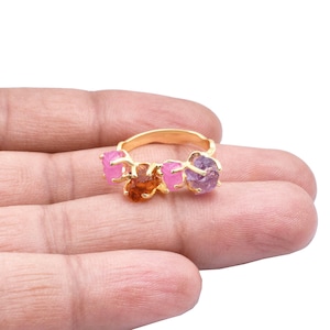 May include: A gold-toned ring featuring a cluster of raw gemstones. The ring showcases a central orange-brown stone flanked by two pink and one purple stone. The gemstones are irregularly shaped, adding a natural, organic aesthetic to the jewelry.