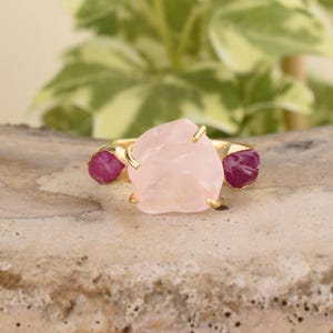 Può includere: Anello in oro con una grande pietra di quarzo rosa grezza rosa e due piccole pietre di rubino rosse su entrambi i lati.
