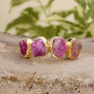 May include: A gold ring with four pink gemstones set in a bezel setting. The gemstones are irregular in shape and have a rough, natural look.
