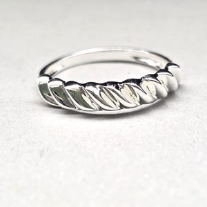 Puede incluir: Un anillo de plata con un diseño de cuerda trenzada. El anillo está hecho de un metal brillante y reflectante, con un acabado liso y pulido. El patrón trenzado envuelve toda la banda del anillo.