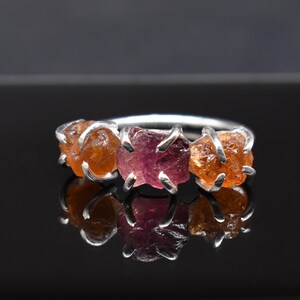 May include: A silver ring with three raw gemstones. The center stone is pink and the two outer stones are orange.