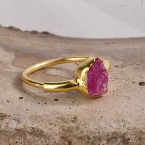 May include: A gold ring with a raw pink ruby gemstone. The ring is simple and elegant, with a smooth band and a natural-looking stone.