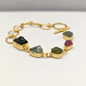 May include: Gold-toned bracelet featuring a series of colorful raw gemstones. The bracelet includes black, teal, green, and pink stones, each set in a gold bezel. The clasp is a simple gold toggle. A unique piece of jewelry.
