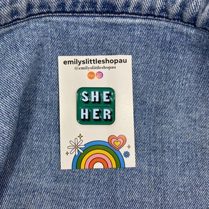 May include: Green enamel pin with the words "SHE HER" in white letters. The pin is on a white card with a rainbow and a daisy.