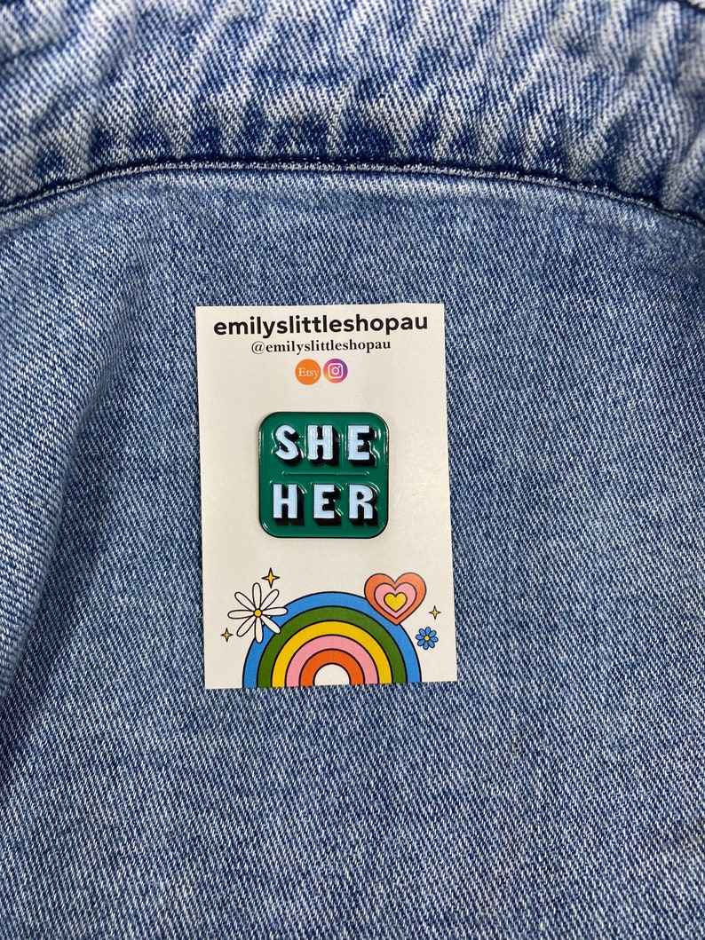 May include: Green enamel pin with the words "SHE HER" in white letters. The pin is on a white card with a rainbow and a daisy.
