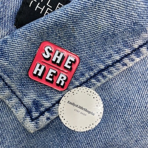 May include: A pink enamel pin with the words "SHE HER" in white letters. The pin is on a blue denim jacket with a white sticker that says "EmilysLittleShopAu ETSY SHOP".