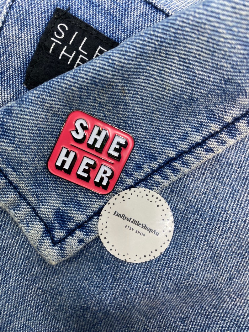 May include: A pink enamel pin with the words "SHE HER" in white letters. The pin is on a blue denim jacket with a white sticker that says "EmilysLittleShopAu ETSY SHOP".