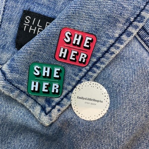 May include: Two enamel pins with the words "She Her" in white letters on a green and pink background. The pins are on a blue denim jacket. A white sticker with the text "EmilysLittleShopAu ETSY SHOP" is also on the jacket.