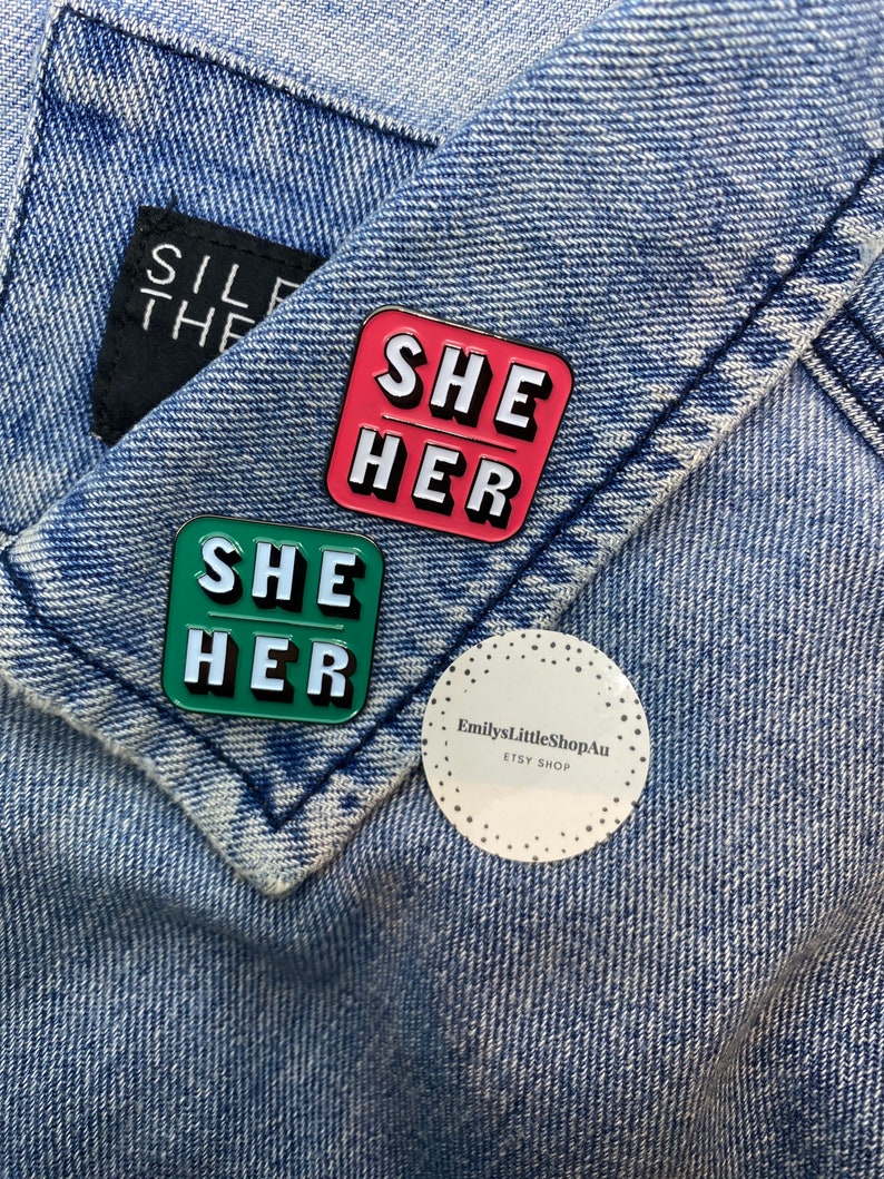May include: Two enamel pins with the words "She Her" in white letters on a green and pink background. The pins are on a blue denim jacket. A white sticker with the text "EmilysLittleShopAu ETSY SHOP" is also on the jacket.