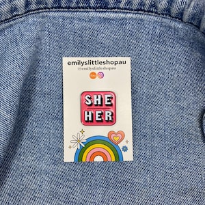 May include: Pink enamel pin with the words "SHE HER" in white letters. The pin is on a white card with a rainbow and a daisy.