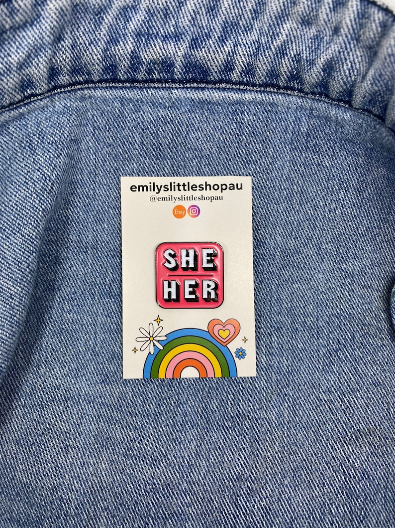 May include: Pink enamel pin with the words "SHE HER" in white letters. The pin is on a white card with a rainbow and a daisy.