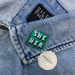 May include: Green enamel pin with the words "SHE HER" in white letters. The pin is on a blue denim jacket.