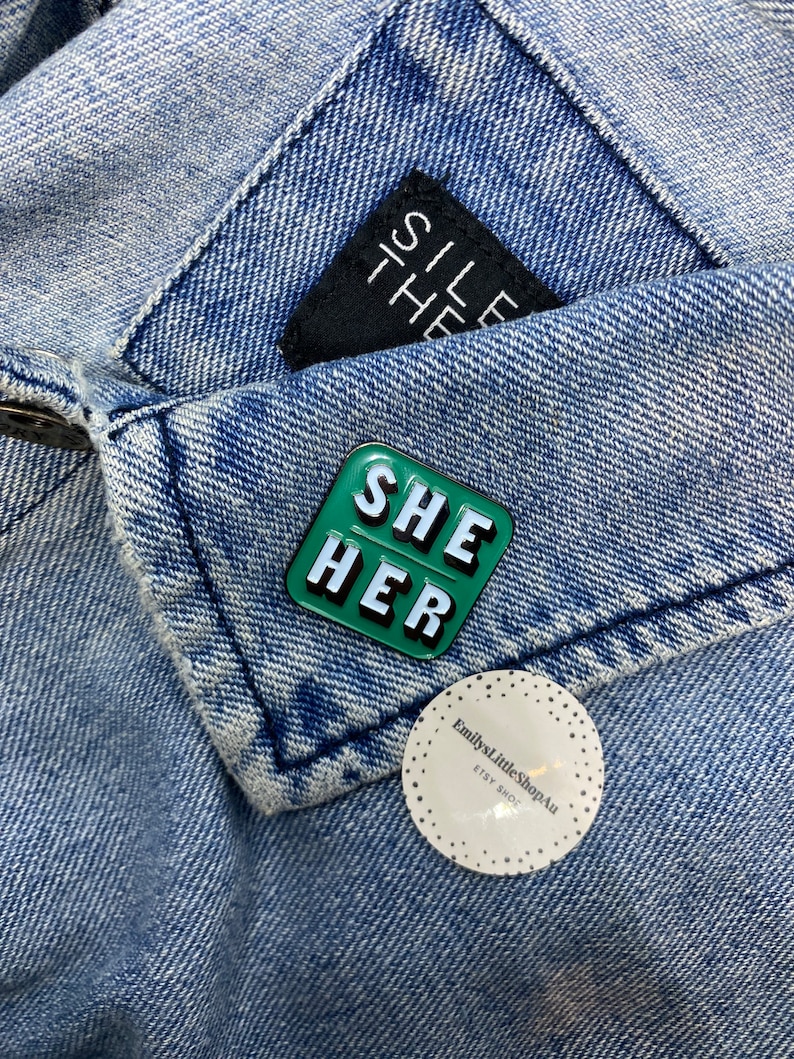 May include: Green enamel pin with the words "SHE HER" in white letters. The pin is on a blue denim jacket.