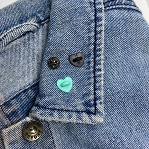 May include: A blue denim jacket with a button closure and a small blue heart pin on the collar. The jacket has a label that says "Theory" on the button.
