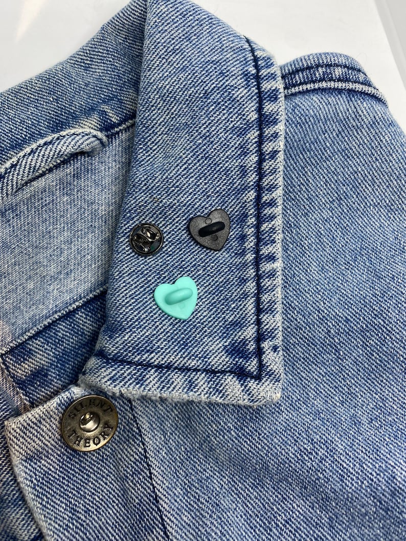 May include: A blue denim jacket with a button closure and a small blue heart pin on the collar. The jacket has a label that says "Theory" on the button.