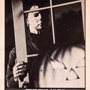 Könnte beinhalten: Ein Schwarzweiß-Plakat, das für den Film "Halloween" wirbt, mit dem Slogan "Trick or Treat Comes One Night Early!" Das Plakat zeigt eine Nahaufnahme von Michael Myers, dem Antagonisten des Films, der durch ein Fenster späht. Das Plakat enthält auch den Text "First Time on TV!" und die Uhrzeit "8:00PM".