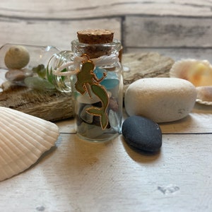 May include: A small glass bottle filled with sea glass and a mermaid charm. The bottle has a cork stopper and is sitting on a piece of driftwood with seashells and stones.