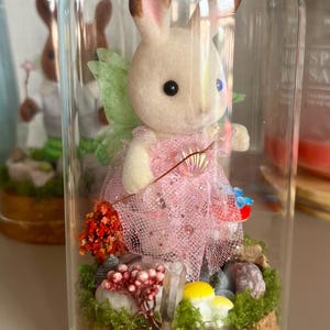 May include: A fairy bunny figurine in a glass dome. The bunny wears a pink dress and green wings, surrounded by miniature flowers, mushrooms, and moss. The dome rests on a cork base, creating a miniature scene.