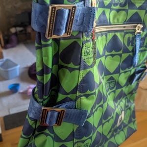 May include: Green and navy tote bag with a heart pattern. The bag has blue straps and metal buckles. Text in Japanese is visible on the bag. The bag has a zipper closure and multiple pockets.