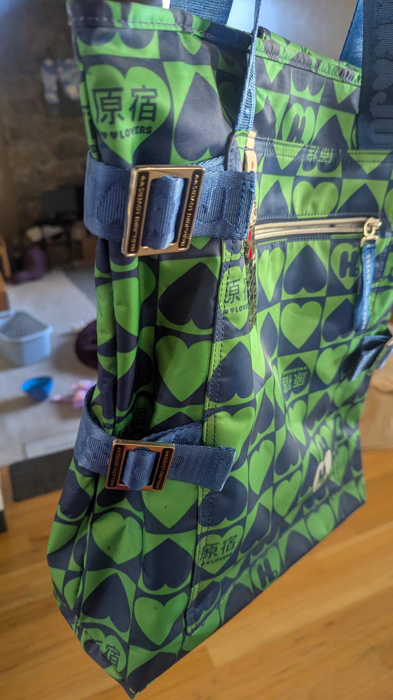 May include: Green and navy tote bag with a heart pattern. The bag has blue straps and metal buckles. Text in Japanese is visible on the bag. The bag has a zipper closure and multiple pockets.