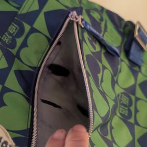May include: Green and blue tote bag with a heart and square pattern. The bag has a zipper pocket and blue straps. The bag has a tag that says "LOVERS".