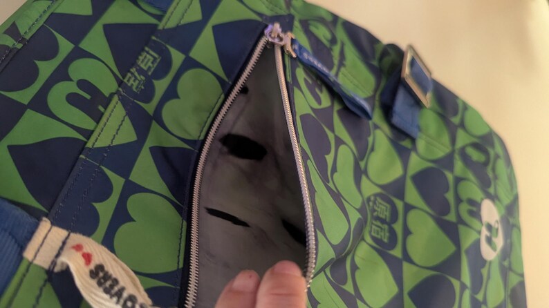 May include: Green and blue tote bag with a heart and square pattern. The bag has a zipper pocket and blue straps. The bag has a tag that says "LOVERS".