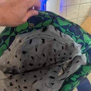 May include: A tote bag with a green and navy blue floral pattern. The bag has a blue strap and a gray interior with black spots. The bag is open, revealing the interior and a zipper pocket. The bag is made of a durable material.