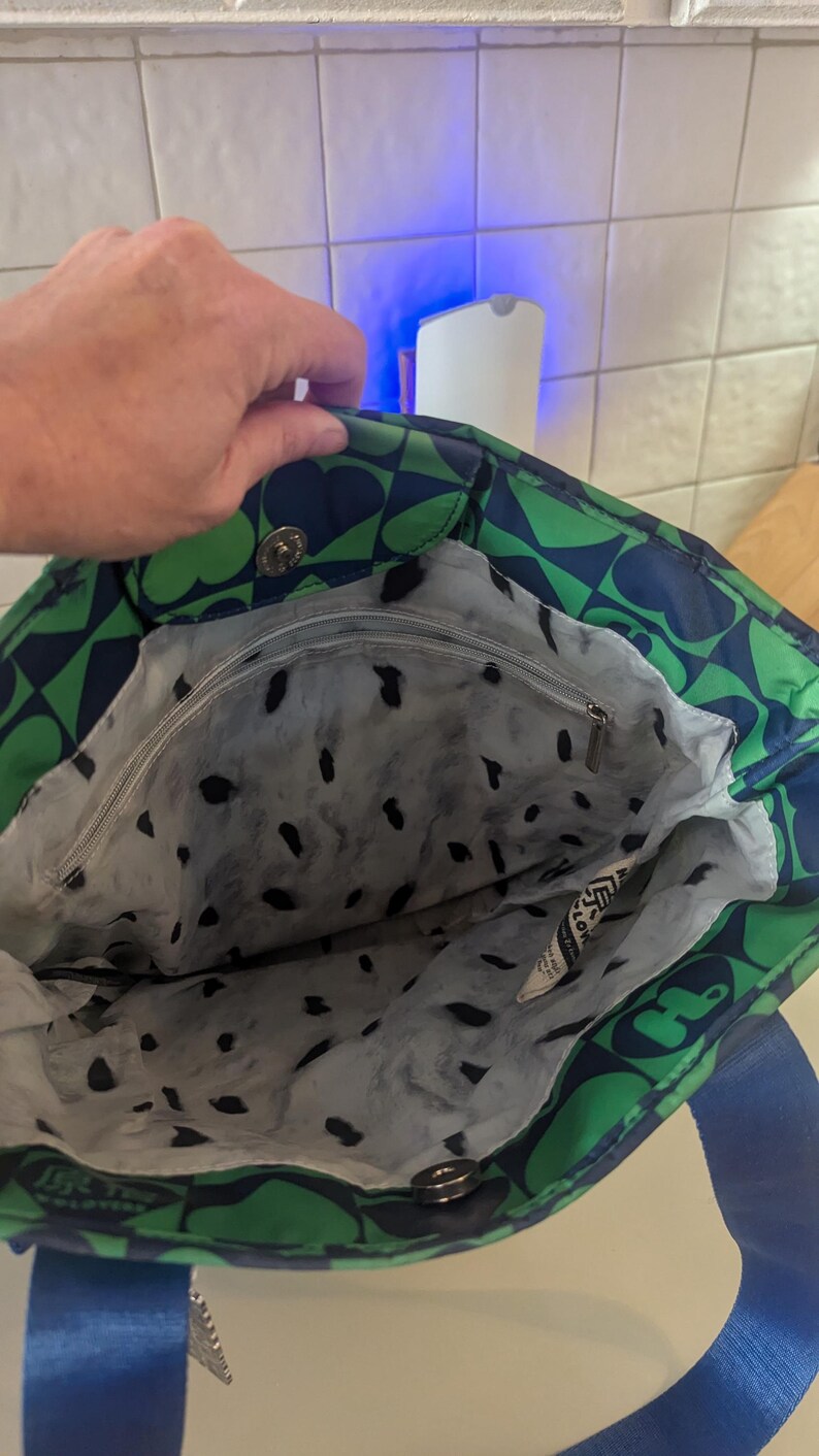 May include: A tote bag with a green and navy blue floral pattern. The bag has a blue strap and a gray interior with black spots. The bag is open, revealing the interior and a zipper pocket. The bag is made of a durable material.