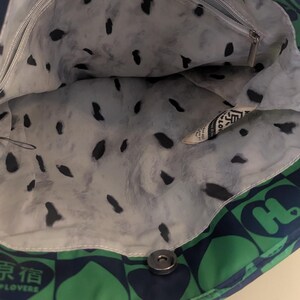 May include: Open tote bag with a green and navy blue heart pattern on the exterior. The interior lining is white with black splotches. A small zippered pocket is visible inside. The bag is closed with a silver snap.