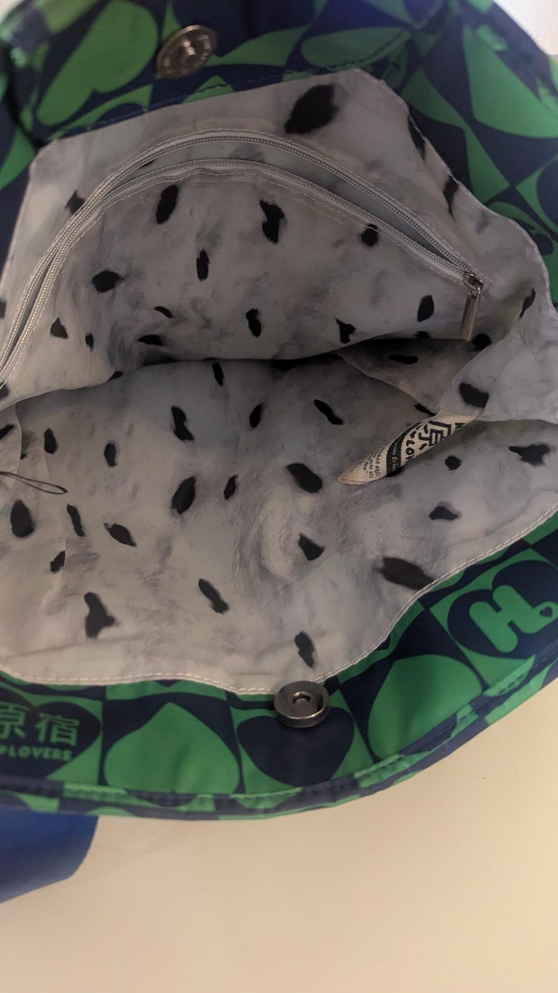 May include: Open tote bag with a green and navy blue heart pattern on the exterior. The interior lining is white with black splotches. A small zippered pocket is visible inside. The bag is closed with a silver snap.