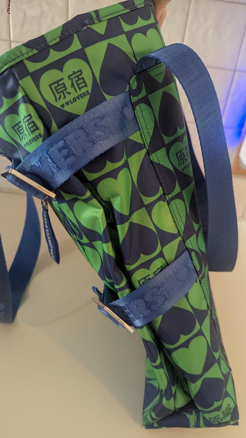 May include: A green and navy tote bag with a heart pattern. The bag has blue straps and the text "原宿" and "LOVERS" in white. The bag is made of a shiny material and has silver hardware.