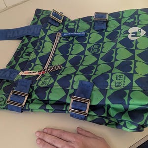 May include: A green and blue tote bag with a heart pattern. The bag has blue straps and buckles. The text "HARJUKU LOVERS" is printed on the strap. A zipper is visible on the front of the bag.