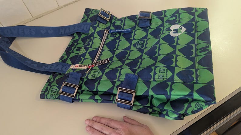 May include: A green and blue tote bag with a heart pattern. The bag has blue straps and buckles. The text "HARJUKU LOVERS" is printed on the strap. A zipper is visible on the front of the bag.
