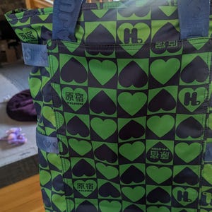 May include: Green and navy tote bag with a repeating heart pattern. The bag features a navy blue strap and the text "原宿" and "W LOVERS" in white. The bag has a square shape.